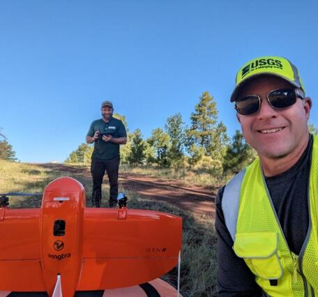 two scientists in USGS gear look into a camera, one next to a UAS and one behind it holding remote control