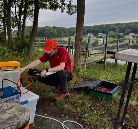 A man wearing a red shirt and orange hat crouches down in front of a peristaltic pump in front of a dock.