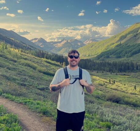 Male with brown hair in a grey shirt, black shorts, and glasses smiles amidst stunning mountain scenery