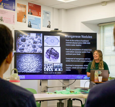 A women stands beside a large presentation screen in front of a classroom.