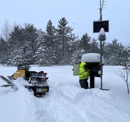 A Hydrologic Technician works on equipment inside a monitoring location housing in knee-deep snow next to a snowmobile