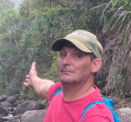 man with a green hat, red shirt, and blue backpack gestures to tropical forest in background