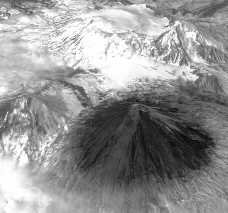 Satellite image of several large mountains, most snow-covered except the foreground volcano that's dark and covered in ash
