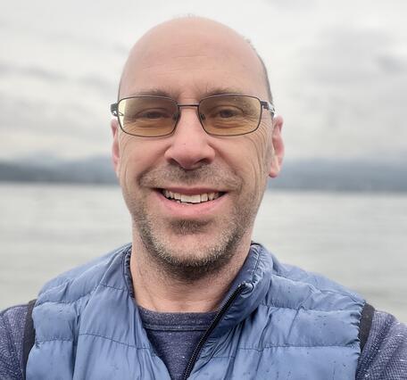 Man with blue vest and blue shirt and glasses smiling 