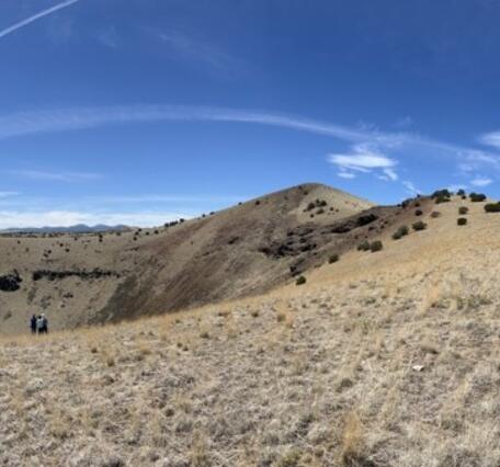 A panoramic photo shows a deep pit with layered crater walls and gentle outer slopes. The walls of the crater are rocky, but the rest of the slopes are covered in gray-tan grass and dark green shrubs. At right, a large SUV is parked on one slope. 