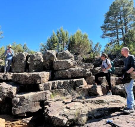 In this photo, three scientists are examining a large pile of slabs of lava. One man wearing a blue button-down shirt and khaki hat is perched on an upper level of the slabs at left, a young woman with braided hair has one foot perched on a slab like a certain pirate at middle right, and a tall bald man in a black shirt stands on top of a pavement of hexagonal lava columns at far right. In the background, scrubby pine trees and bushes grow atop the lava. 