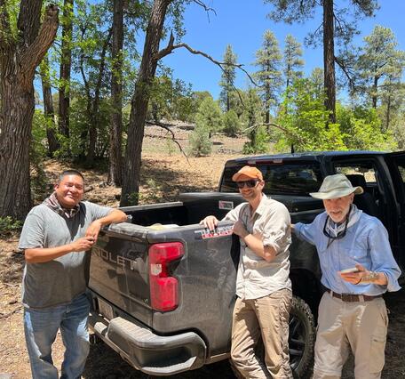 A photograph of three men leaning against the bed of a dusty black truck. The man on the left has tan-colored skin, short dark hair, and is wearing dusty jeans, a gray t-shirt, and a colorful bandanna. The man in the center is wearing khaki pants, a light blue button-down shirt, an orange USGS ballcap, and dark sunglasses. The man on the right is older and wearing khaki pants, a blue button-down shirt, a white bandanna, and a khaki hat, with sunglasses on a lanyard around his neck. 