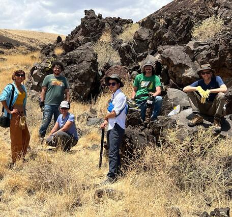 In this photo, a group of six people stand and sit on the edge of a lava flow made up of craggy black boulders. From left to right, a woman wearing brown pants, blue and yellow shirts, and a kerchief; a bearded man wearing jeans and a green t-shirt; a kneeling woman wearing black pants, a blue t-shirt and a khaki ballcap; a man wearing black pants, a white shirt and a gray hat; a man with a moustache wearing jeans, a green t-shirt and a gray ballcap; and a sitting man wearing khaki pants and a blue shirt.