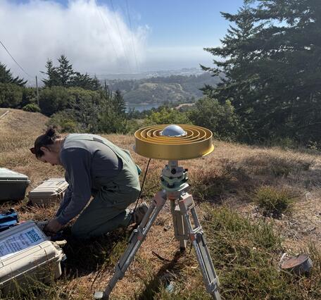 USGS scientist kneels beside gold ring GNSS antenna on a hillside above San Andreas Lake with fog along the distant coast.