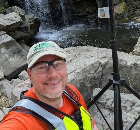 Scientist standing next to air sampling equipment with waterfall in background