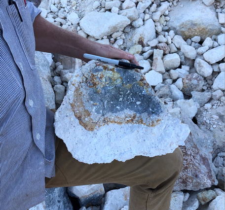 A geologist in a blue shirt and khakis supports a basketball-sized chunk of lava on his knee. A bright white "rind" makes up about half of the chunk, surrounding a core of dark gray lava speckled with white crystals. A marker rests on the chunk for a sense of scale, and similar white chunks of lava make up the background of the photo. 