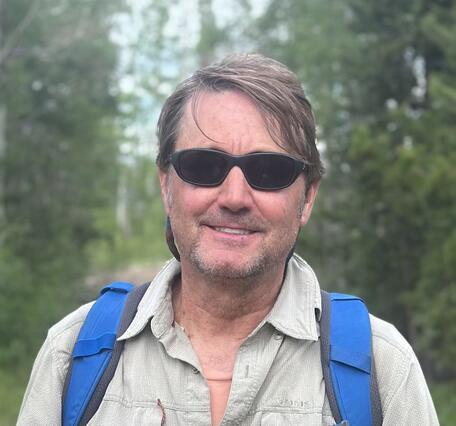 photo of a man's head, man wearing sunglasses and blue backpack straps, trees in the background