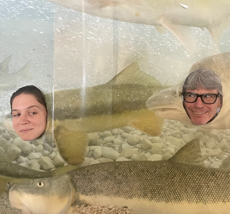 Two USGS scientists pose behind a humpback chub cutout display, their faces framed where the fish’s heads would be.