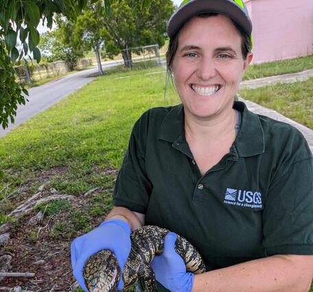 a woman in a dark green shirt and purple gloves holds a black and white lizard, pink building, grass, and trees behind