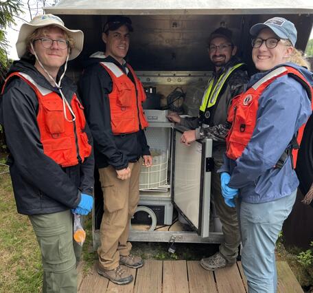 Four smiling scientists stand near an metal housing for an ISCO sampler
