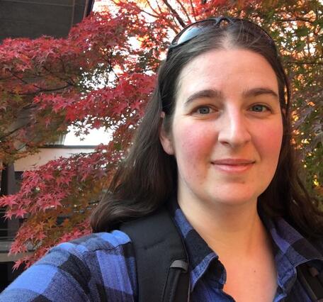 Woman with long brown hair and sunglasses, wearing a flannel shirt. 
