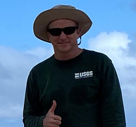man in long sleeve green USGS shirt and wide brimmed tan hat and sun glasses give the hang loose sign. 