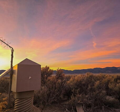 Yellow-orange sunset over desert landscape with groundwater monitoring station and distant mountains.
