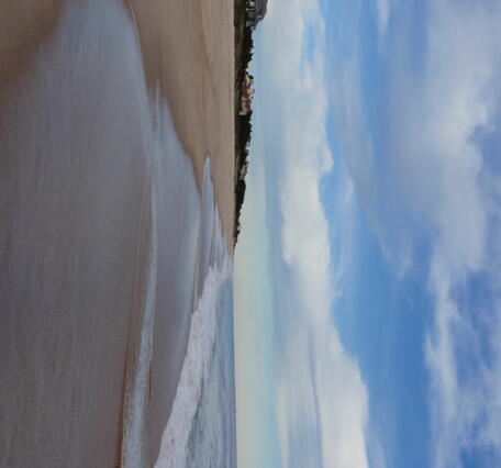 Calm ocean waves on a beach with houses and a cloudy sky in the background