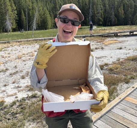 Smiling woman standing on a boardwalk and holding an open pizza box. Scrubby ground and forested hill in background.