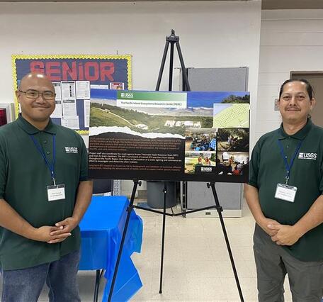 Two people in green shirts stand on either side of a poster inside a community center room.