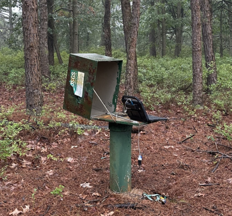 A computer attached to a probe going down a pipe inside a steel box in the woods.