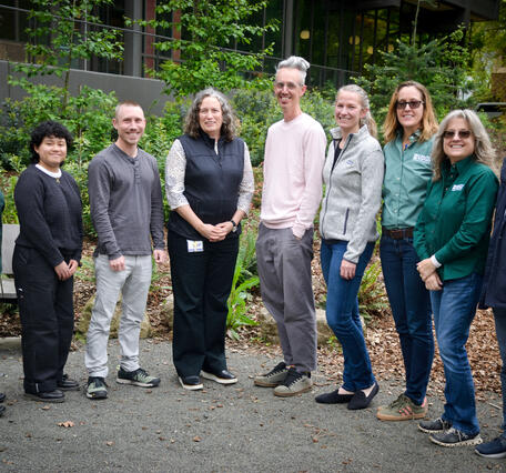 9 people stand in a line with the tallest people in the middle. They are professionally dressed outside amongst greenery