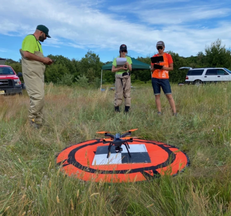 three people in a grassy field stand around an orange launch pad preparing to deploy a small drone with thermal cameras