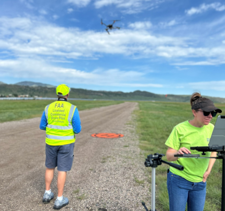 two scientists in bright yellow shirts stand on an airplane landing strip in a field, with a small UAS in the air above them