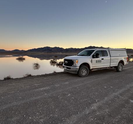 White USGS truck parked on dirt road near shallow-water playa at dusk with silhouetted mountains.