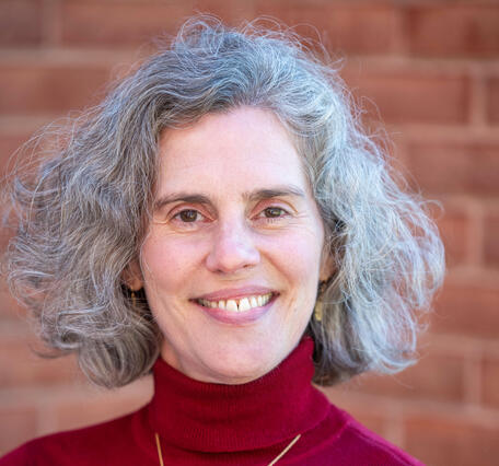 Headshot of a woman with chin-length curly grey hair wearing a maroon turtleneck in front of a brick wall.