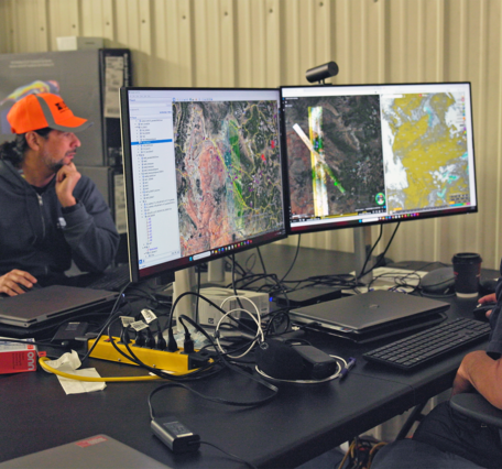 Two people in a hangar at a workstation displaying hyperspectral survey maps, surrounded by laptops and field equipment