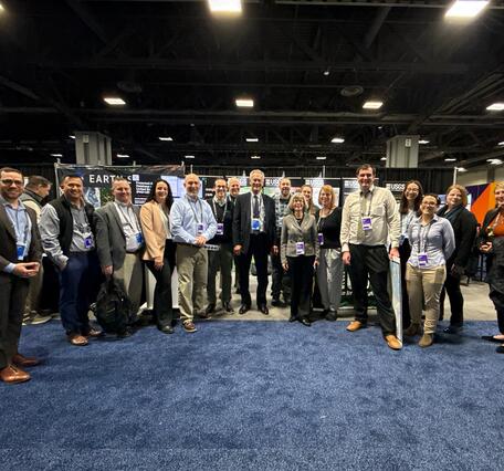 17 people stand in a row at a conference, smiling for the camera