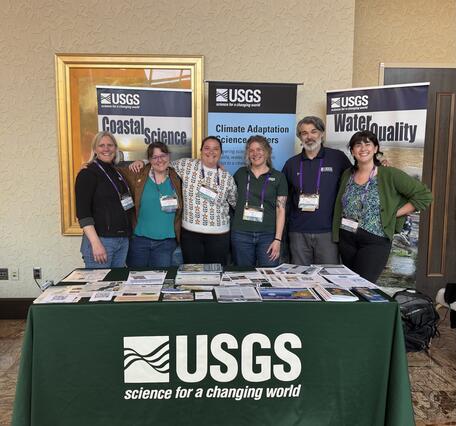 Six people stand at behind a table covered in a tablecloth that has the USGS logo on it.