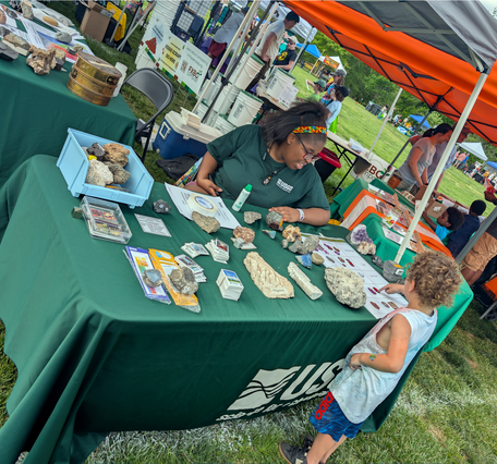 A USGS employee sits behind an outreach table during an outdoor event, talking with a child about the materials on the table