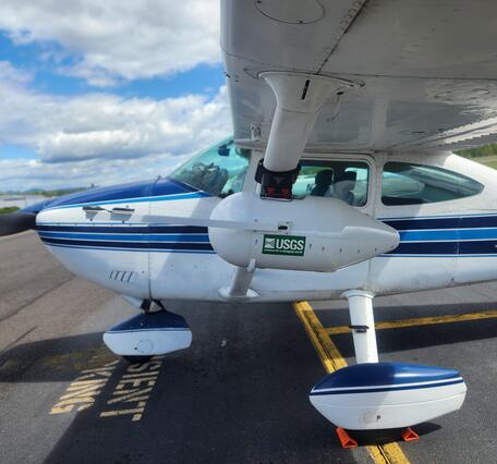 a small white plane with a blue stripe down the middle. USGS sticker on a white enclosure housing Multispectral camera system
