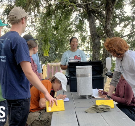 students gather over grey picnic table to look at equipment while note taking in yellow field notebooks