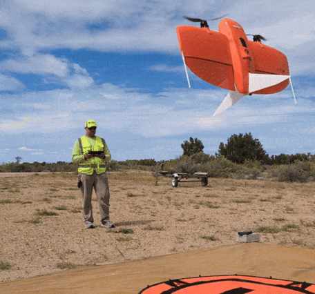 A man in neon yellow gear holds a remote control, an orange drone rises up and lands on a helipad in front of the man
