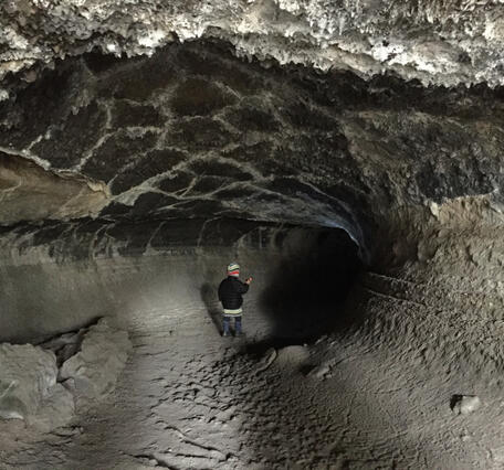 A small child wearing warm winter clothing is viewed from the back, standing in a natural tunnel of dark volcanic rock. The roof of the tunnel is crusted with white minerals and drips of hardened rock, and to the right horizontal raised lines follow the wall of the tunnel to show lava flow levels.