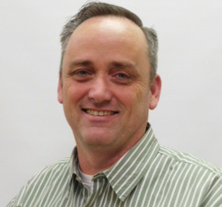 Photo of Vincent Delaney, dark hair with grey highlights, smiling, and wearing a collared shirt with green vertical pin stripes.