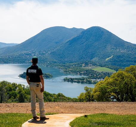 A geologist wearing a green USGS shirt and ballcap is seen from the back, standing at the top of a hill overlooking a lake and a large mountain. The lake is divided by a long peninsula dotted with homes and trees, and the mountain has two prominent peaks that are covered in dark forest.