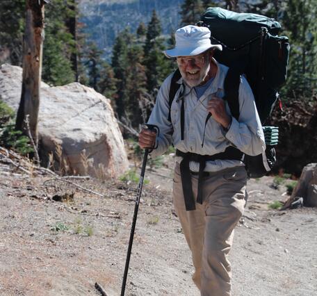 An older geologist wearing khakis, a light blue shirt, and a fisherman's hat hikes up a mountain carrying a large backpack and holding a hiking pole. He is smiling at the photographer.