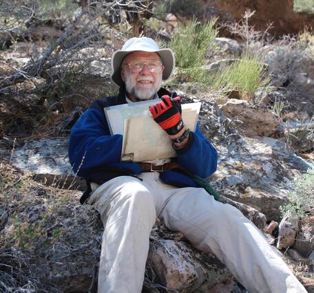 A man with white hair and a beard, wearing khakis, a blue shirt, and a fisherman's hat, reclines on a rock outcrop. He is holding a bundle of geologic maps and smiling.
