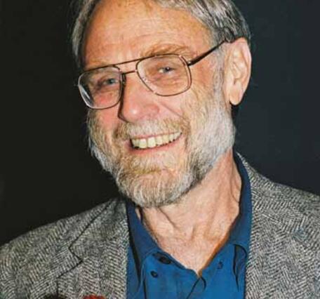 An older man with a salt-and-pepper beard and hair and square glasses, wearing a speckled gray suit and a teal button-down shirt, smiles at the camera. He is holding a red rose. 
