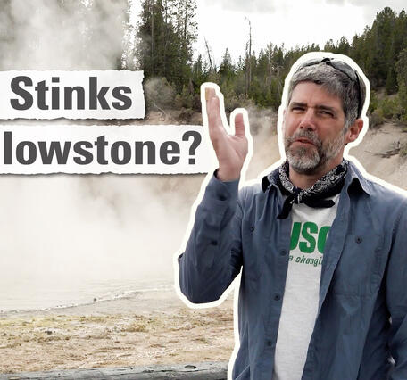 Man in blue shirt waves his hand in front of his face and stands in front of a boiling hot spring