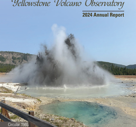 Muddy eruption from a blue water spring on barren ground, forested hills and blue sky in the background