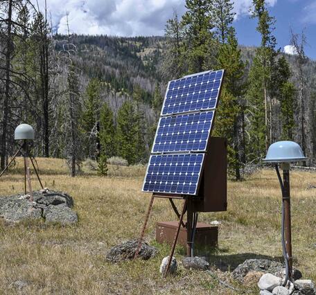 GPS antenna with gray dome on rock in middle ground, solar panels and satellite antenna in foreground, in grassy meadow