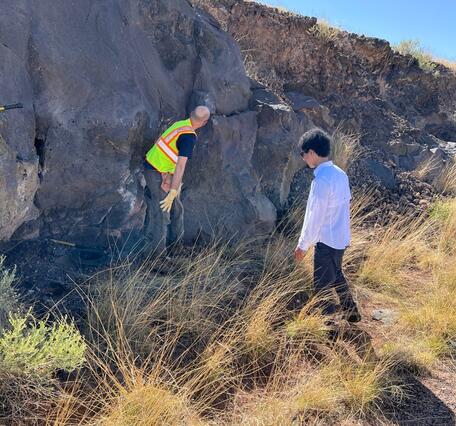 Two men examine a vertical outcrop of gray rock about twice their height, with brown grass at their feet and blue sky above