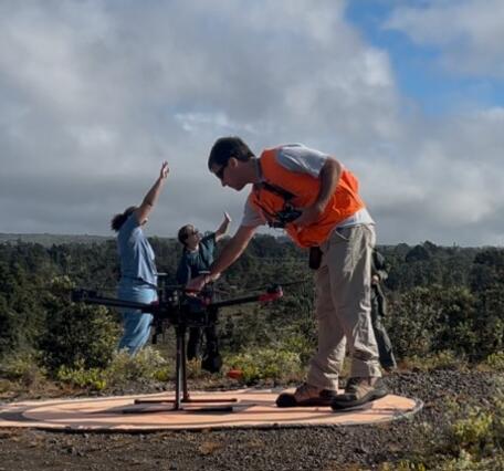 Color photo of a USGS scientist in an orange vest sitting up a UAS while two scientists in the background observe for birds