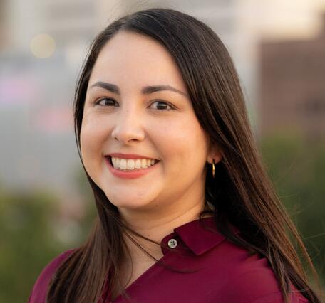 Color photo of Zulimar smiling. She has long brown straight hair and is wearing a marroon shirt with city buildings in the background. 